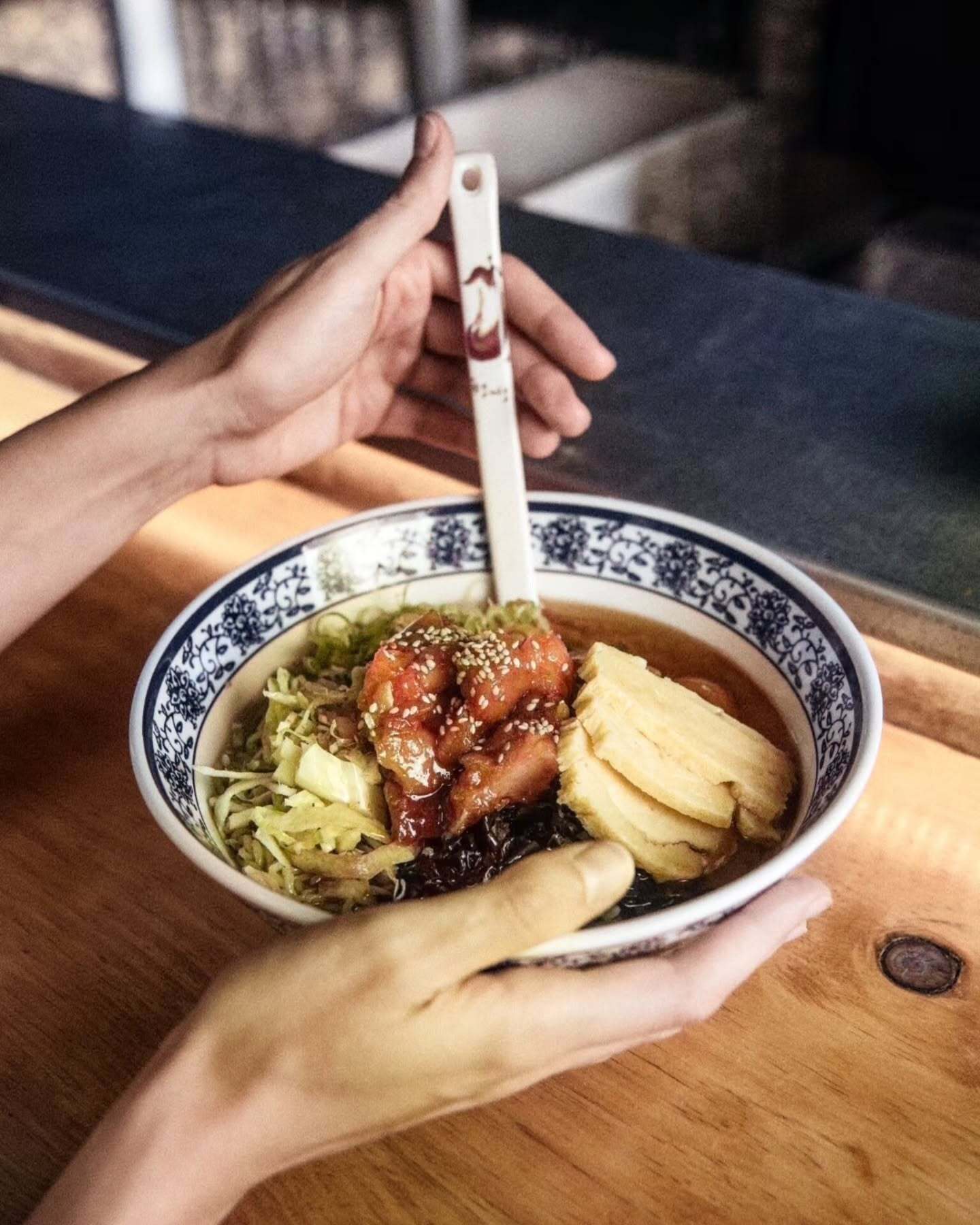 Manos sosteniendo un bowl de ramen con chashu, tofu y col